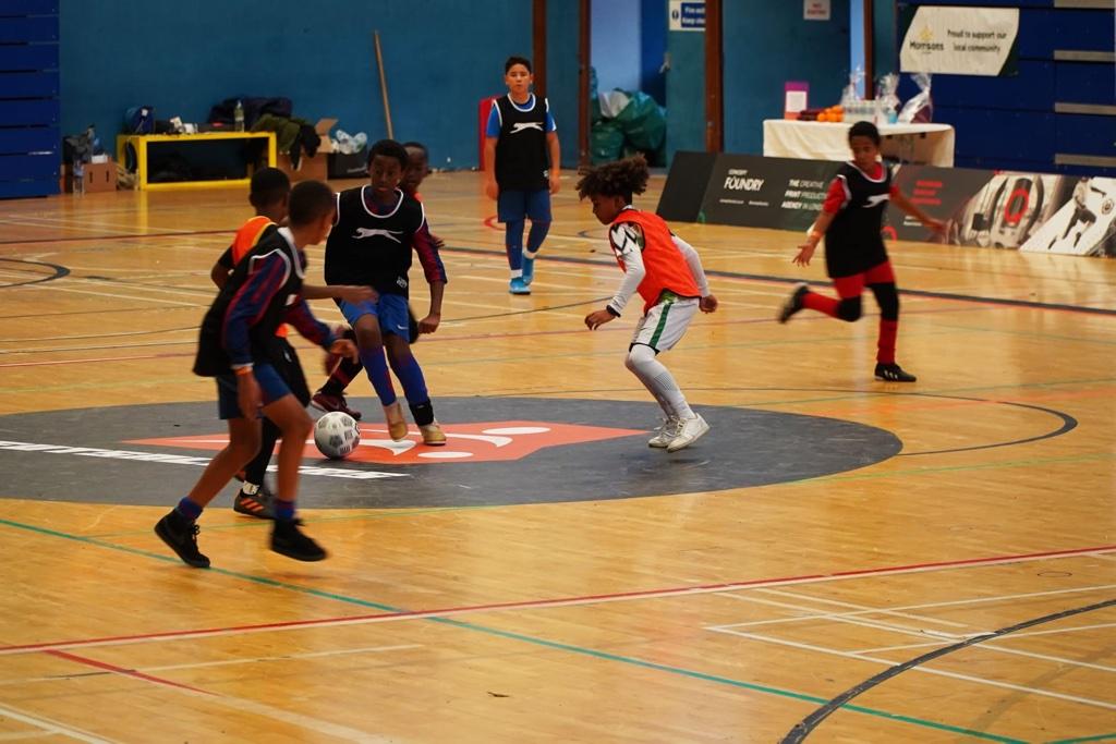 A group of young children playing soccer on a court, with basketball playing in the background.